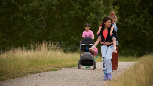 Two children walk along a gravel path through tall grass, with one child carrying a smaller child on their back. Behind them, another child pushes a stroller along the same path. Trees and greenery line the route.